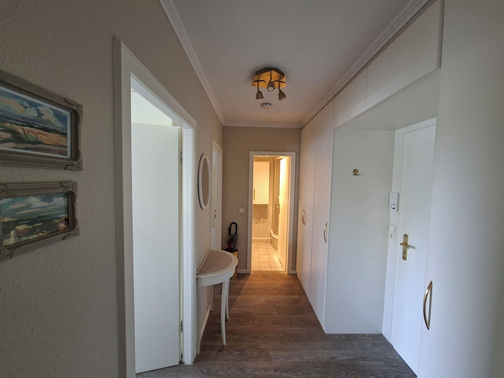 A hallway with white walls, a wooden floor, and a ceiling light fixture. There are two doors on the right, one with a gold handle and a small number above it. A white bench is against the wall on the left, and a framed picture hangs above it.