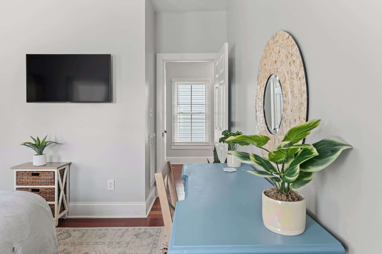 A room with a blue table, a potted plant, a wall-mounted mirror, a flat-screen TV, and a doorway leading to another room.