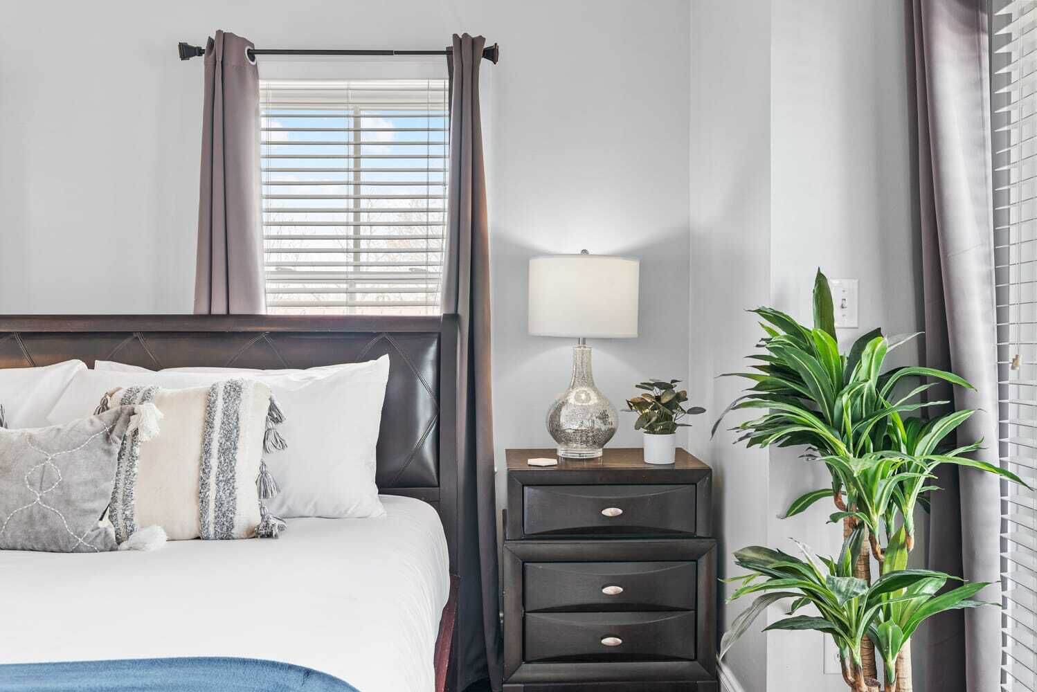Un dormitorio ordenado con una cama, mesitas de noche, una lámpara y una planta en maceta.