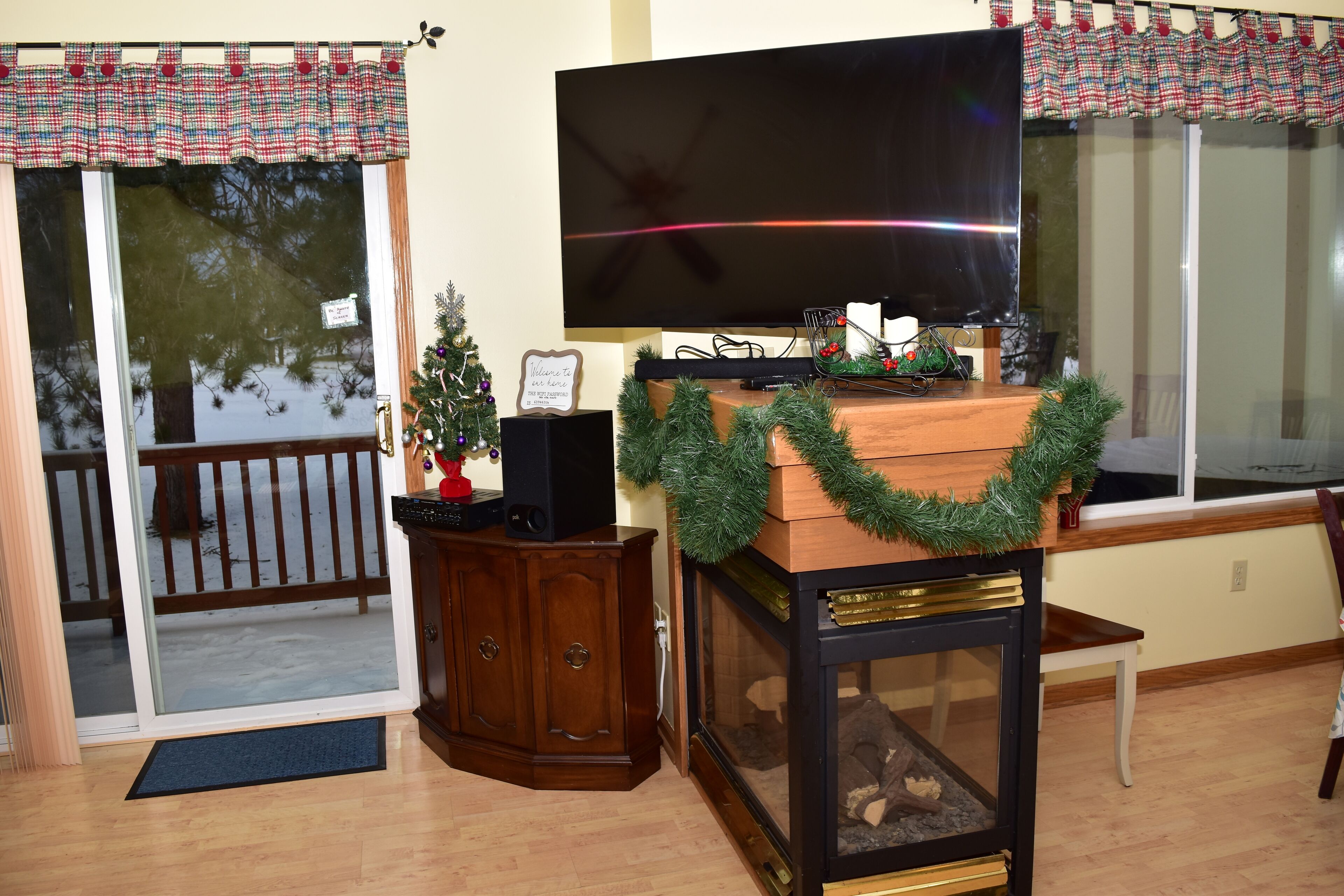 Un salon avec un téléviseur à écran plat posé sur un meuble, une armoire en bois, un petit sapin de Noël et une cheminée avec une étagère.
