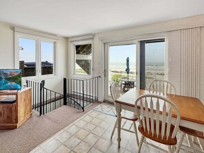 A room with a dining table, chairs, a TV, and a view of the beach through large windows.