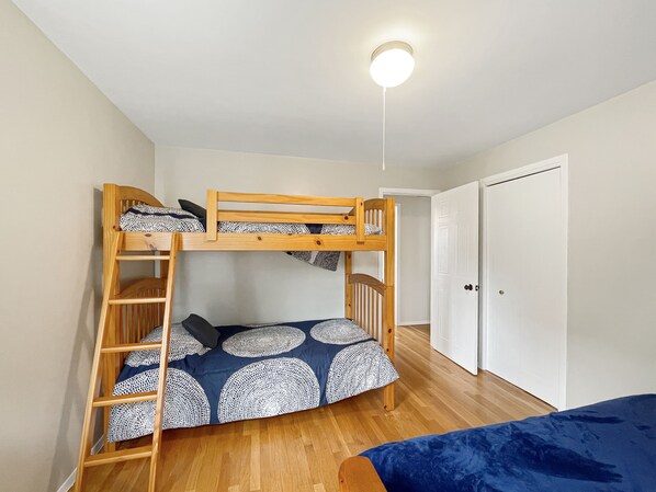 Uma cama de beliche com escada, uma cama de solteiro e uma porta num quarto com piso de madeira.