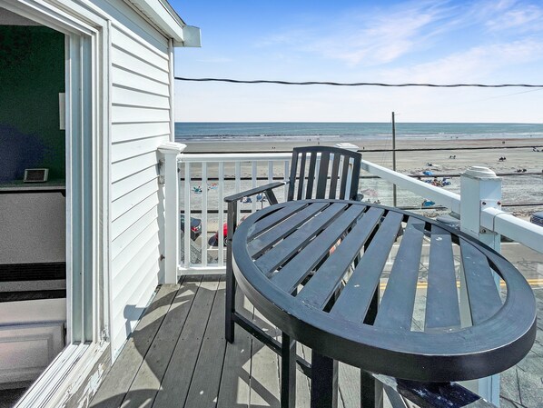 A wooden deck with a round table and chairs, overlooking a beach with people and umbrellas.