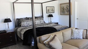 A four-poster bed with a patterned bedspread, a beige sofa with decorative pillows, a nightstand with a lamp, and a framed picture on the wall.