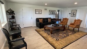 A living room with a black sofa, two armchairs, a coffee table, and a patterned rug. There are framed pictures on the walls and a door in the background.