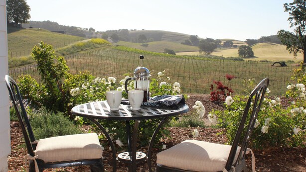 A round metal table with two chairs, a coffee pot, and cups, set in a garden with a view of rolling hills and vineyards.