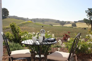 A round metal table with two chairs, a coffee pot, and cups, set in a garden with a view of rolling hills and vineyards.