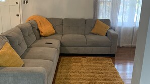Una sala de estar con un sofá seccional gris, dos cojines amarillos, un alfombra de pelo alto marrón, suelo de madera, una puerta blanca y cortinas traslúcidas en una ventana.