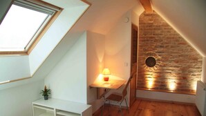 A cozy attic bedroom with a sloped ceiling, a wooden desk with a lamp, a chair, and a small plant. - AN OASIS WHERE TO FIND THE ESSENTIALS<br>AND TAKE A BREAK IN CONSCIOUSNESS & IN (SAINT USUGE)