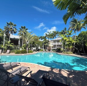 A swimming pool with lounge chairs, surrounded by palm trees and a building with balconies.