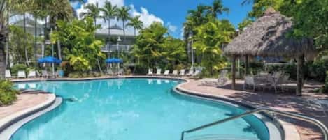 A swimming pool with a curved edge, surrounded by lounge chairs and umbrellas, with palm trees and a thatched-roof structure in the background.