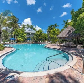 A swimming pool with a curved edge, surrounded by lounge chairs and umbrellas, with palm trees and a thatched-roof structure in the background. - Beautiful Key West Cottage (Key West)
