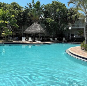 A swimming pool with lounge chairs and a thatched-roof structure surrounded by trees.