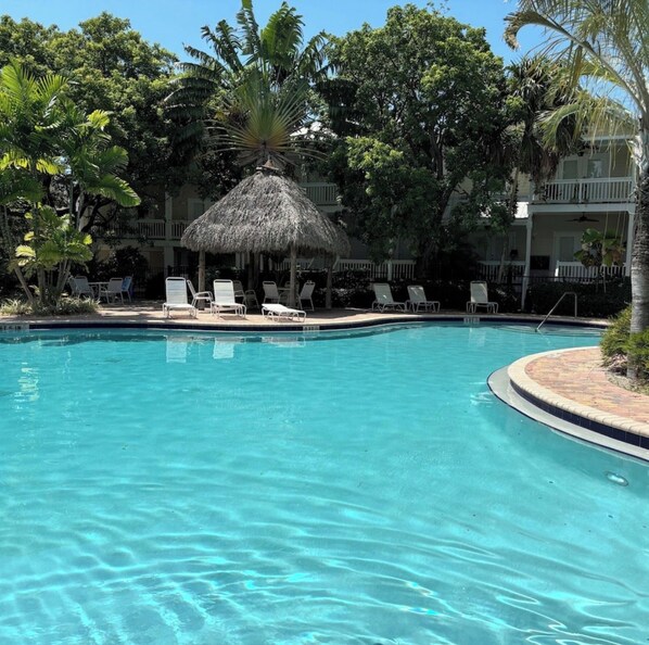 A swimming pool with lounge chairs and a thatched-roof structure surrounded by trees. - Beautiful Key West Cottage (Key West)