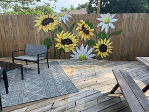 Eine Holzterrasse mit einer rustikalen Bank und einem Stuhl, einem gemusterten Teppich und einem bemalten Zaun mit Sonnenblumen.