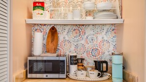 A kitchen counter with a microwave, coffee maker, and a tray with cups. Above the counter, there are shelves with coffee filters, glasses, and dishes. - Diamond head charmer (Honolulu)