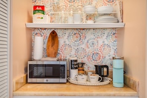 A kitchen counter with a microwave, coffee maker, and a tray with cups. Above the counter, there are shelves with coffee filters, glasses, and dishes.