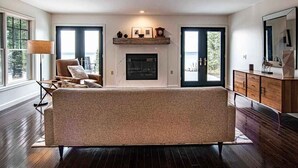 A spacious living room with a large sofa, a fireplace, and a view of the outdoors through the windows. - Sunsets, Sandy Beach and Seclusion; enjoy them all at this stunning lake house (Beaver Island)