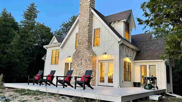 A two-story house with a stone chimney, a covered deck with four chairs, and a large window with yellow curtains. - Sunsets, Sandy Beach and Seclusion; enjoy them all at this stunning lake house (Beaver Island)