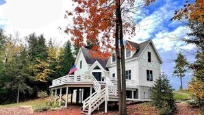 A two-story house with a porch and a flagpole, surrounded by trees and a leaf-covered ground. - Sunsets, Sandy Beach and Seclusion; enjoy them all at this stunning lake house (Beaver Island)