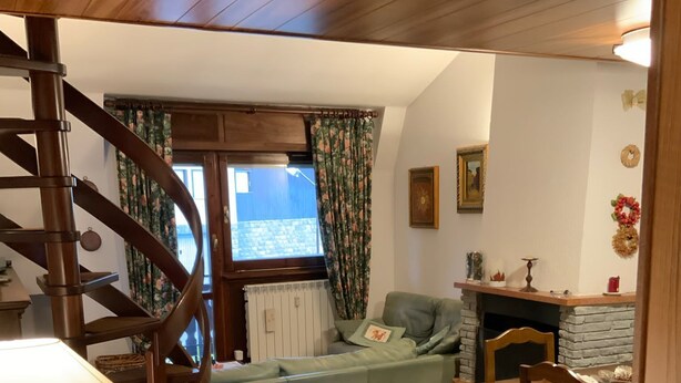 Een gezellige woonkamer met een houten trap, een blauwe bank, een eettafel met stoelen en een open haard.