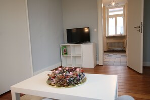Ein Zimmer mit einem Flachbildfernseher auf einer weißen Kommode, einer dekorativen Blumenanordnung auf einem weißen Tisch und Blick auf einen Flur mit Heizkörper und einer Tür, die nach draußen führt.