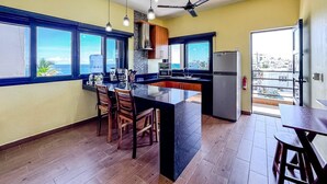 Een moderne keuken met een centraal eiland, houten stoelen, een plafondventilator en grote ramen met uitzicht op de oceaan.