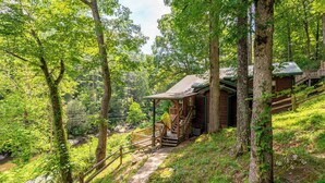 A rustic cabin with a wooden deck and stairs leading to a porch, surrounded by lush greenery and a small stream. - Riverfront • Hot Tub • Convenient Location (Blue Ridge)