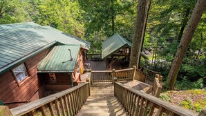 A wooden cabin with a green roof, a wooden deck, and a staircase leading to a covered porch. - Riverfront • Hot Tub • Convenient Location (Blue Ridge)