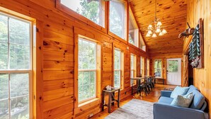 A cozy cabin interior with large windows, a dining area, and a seating area with a blue sofa. - Riverfront • Hot Tub • Convenient Location (Blue Ridge)