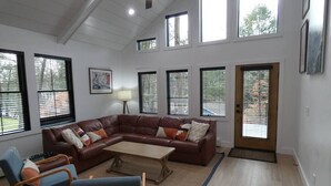 A living room with a brown leather sofa, a wooden coffee table, and a blue armchair. There are multiple windows with blinds, a framed picture on the wall, and a door with a doormat.