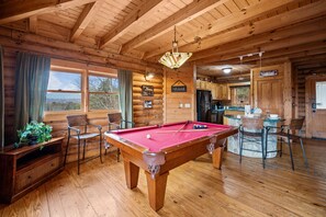 Interior de una cabaña de madera con mesa de billar, sillas, comedor y cocina.
