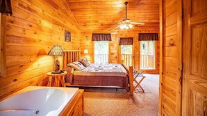 Ein Schlafzimmer in einer Blockhütte mit einem großen Bett, einer Badewanne, einem Deckenventilator und Fenstern mit Vorhängen.