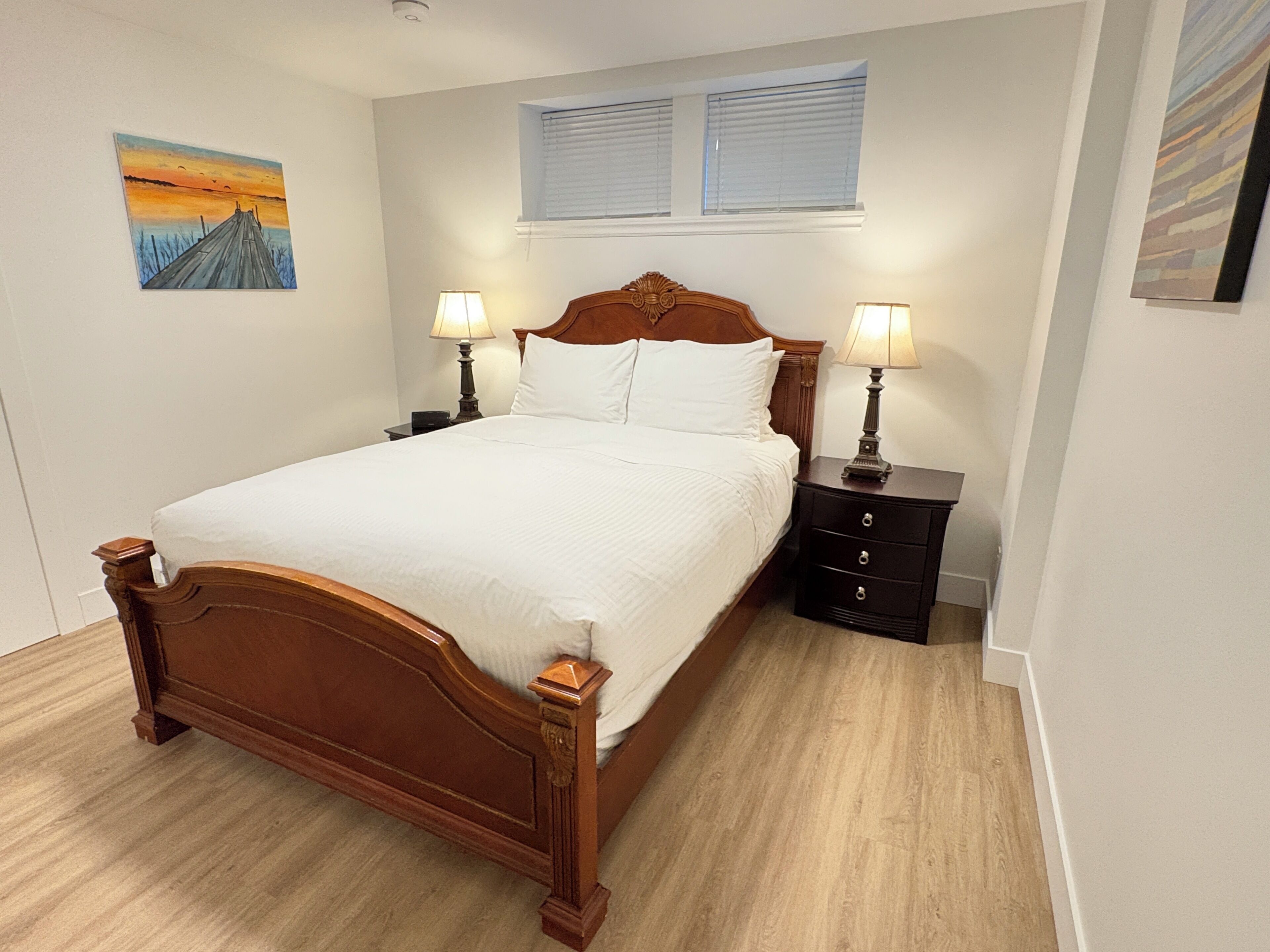 A bedroom with a wooden bed, two bedside tables with lamps, a window with blinds, and a painting on the wall.