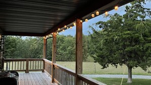 Una terraza de madera con barandilla, luces cálidas y vistas a árboles y césped.