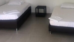 Dos camas individuales con sábanas blancas, una mesita de noche negra pequeña y una pared blanca lisa.