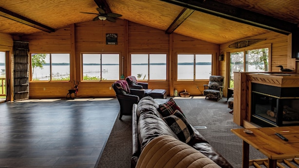A spacious living room with a large window overlooking a lake, a fireplace, and a cozy seating area. - Round Lake Retreat: UP Lakefront Vacation Home (McMillan)