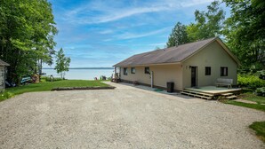 A single-story building with a gravel driveway, a front porch, and a view of a lake. - Round Lake Retreat: UP Lakefront Vacation Home (McMillan)