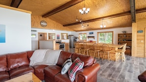 A cozy living room with a leather couch, wooden dining table, and chairs. A kitchen area with wooden cabinets and a refrigerator is visible in the background. - Round Lake Retreat: UP Lakefront Vacation Home (McMillan)