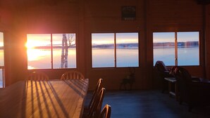 A room with a large wooden table and chairs, a bench, and a view of a sunset over a body of water. - Round Lake Retreat: UP Lakefront Vacation Home (McMillan)