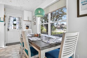 A dining area with a wooden table, blue cushioned chairs, a green pendant light, and a view of trees through large windows.