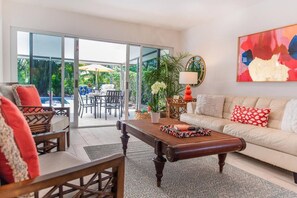 A modern living room with a sofa, coffee table, and chairs. There is a dining area with a table and chairs visible through a glass door leading to a pool. - 606 SeaRenity Resort • Designer Home w/Heated Pool (Naples)