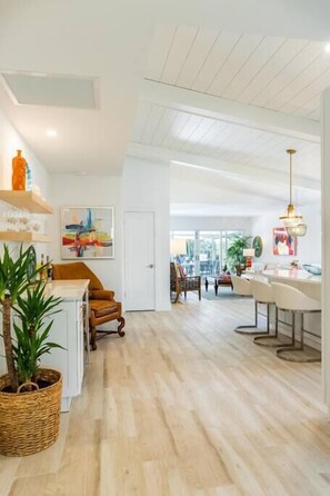A spacious living area with a wooden floor, a dining table, and a kitchen area with a white countertop and a hanging light fixture. - 606 SeaRenity Resort • Designer Home w/Heated Pool (Naples)