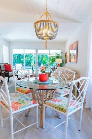 Een eethoek met een glazen tafel, omringd door witte stoelen met bloemkussens, een hangende kroonluchter en uitzicht op het buiten.