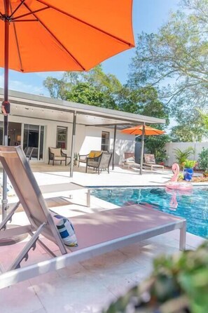 Zwembadgedeelte met loungebedden en parasols, een huis op de achtergrond, en een roze flamingo drijvend in het water.