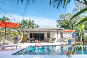 A pool with a pink flamingo float, surrounded by lounge chairs and umbrellas, with a covered patio area and palm trees in the background.