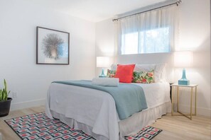 A bedroom with a bed, two bedside tables, a potted plant, a patterned rug, and a framed picture on the wall.