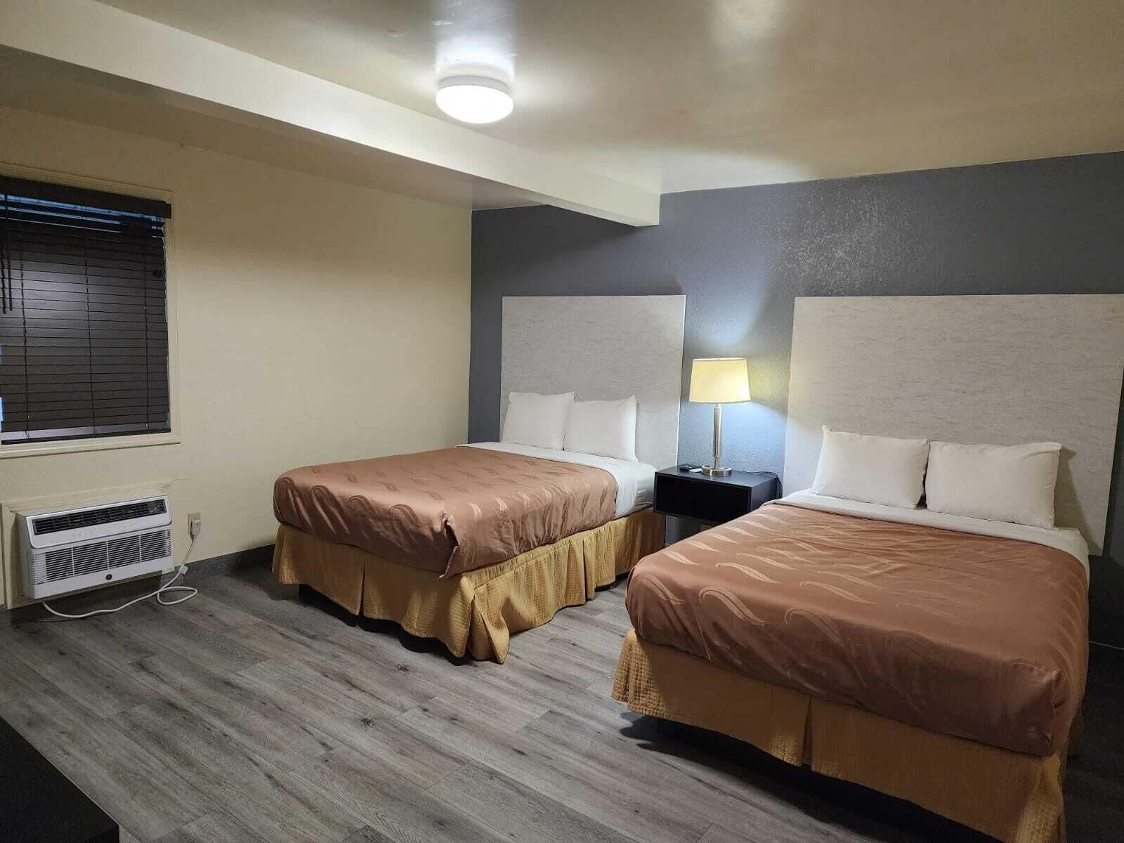 A hotel room with two beds, a nightstand with a lamp, a window with blinds, and an air conditioning unit.
