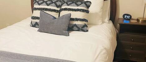 A neatly made bed with white bedding and patterned pillows, a dark wooden headboard, a bedside table with a clock, and a patterned rug.