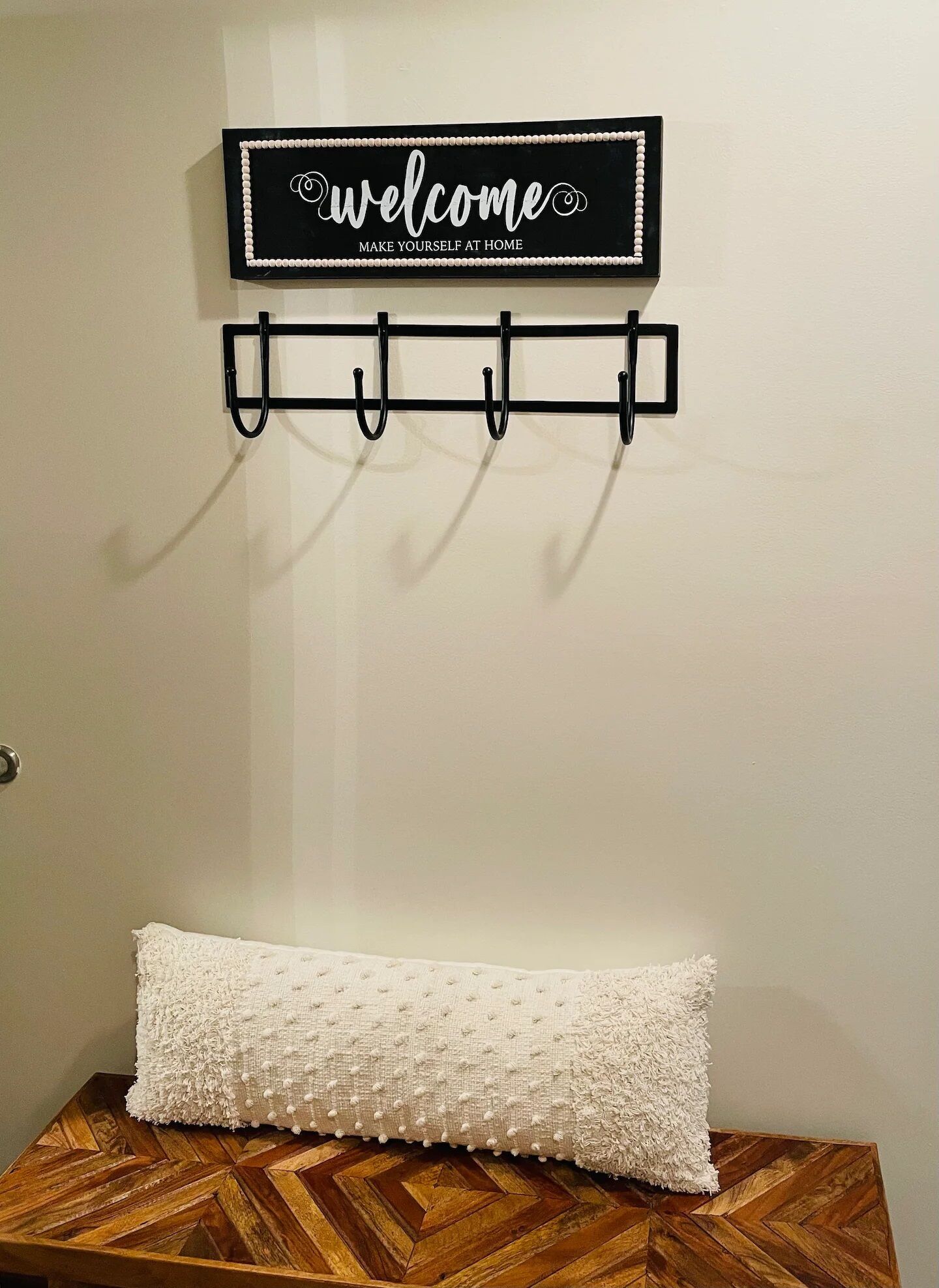 Un letrero montado en la pared con la inscripción 'welcome make yourself at home' sobre un perchero negro fijado a la pared con ganchos. Abajo, un cojín blanco con textura descansa sobre una superficie de madera.